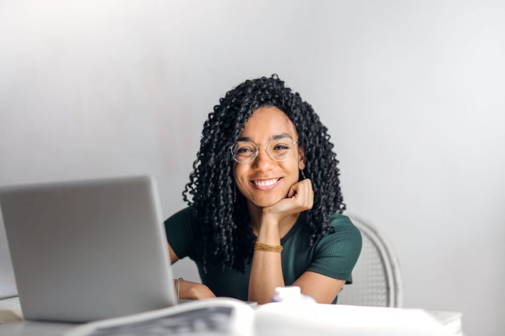 Crafting Captivating Headlines: Your awesome post title goes here Joyful businesswoman with curly hair smiling at camera while using laptop indoors.